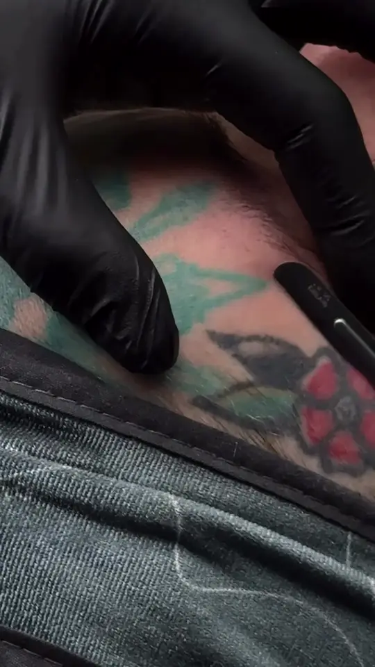 Black-gloved hands dermaplane a client’s cheek, over her floral facial tattoos.