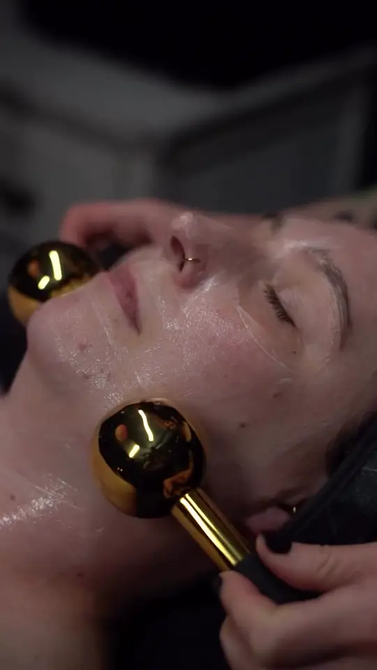Gold globes glide across a client’s cheek in the dark treatment room.